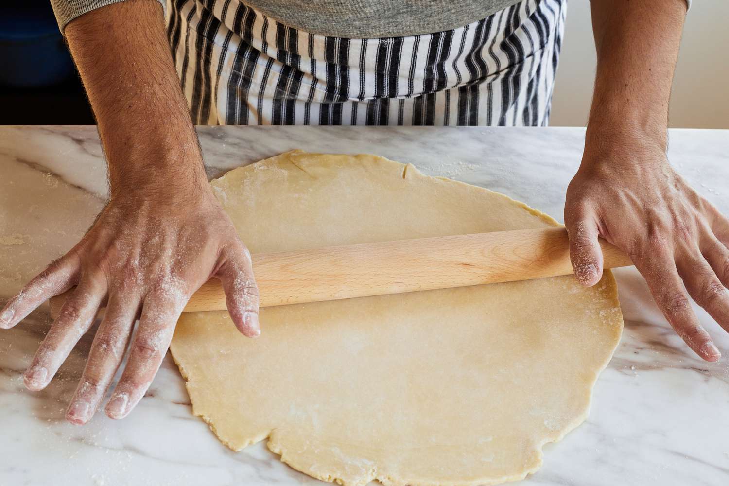 Two hands using a tapered rolling pin to roll out pie crust on a marble surface
