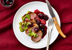 Sliced roasted pork tenderloin with green sauce and roasted cherries on a plate, placed on a red tablecloth with a fork and knife