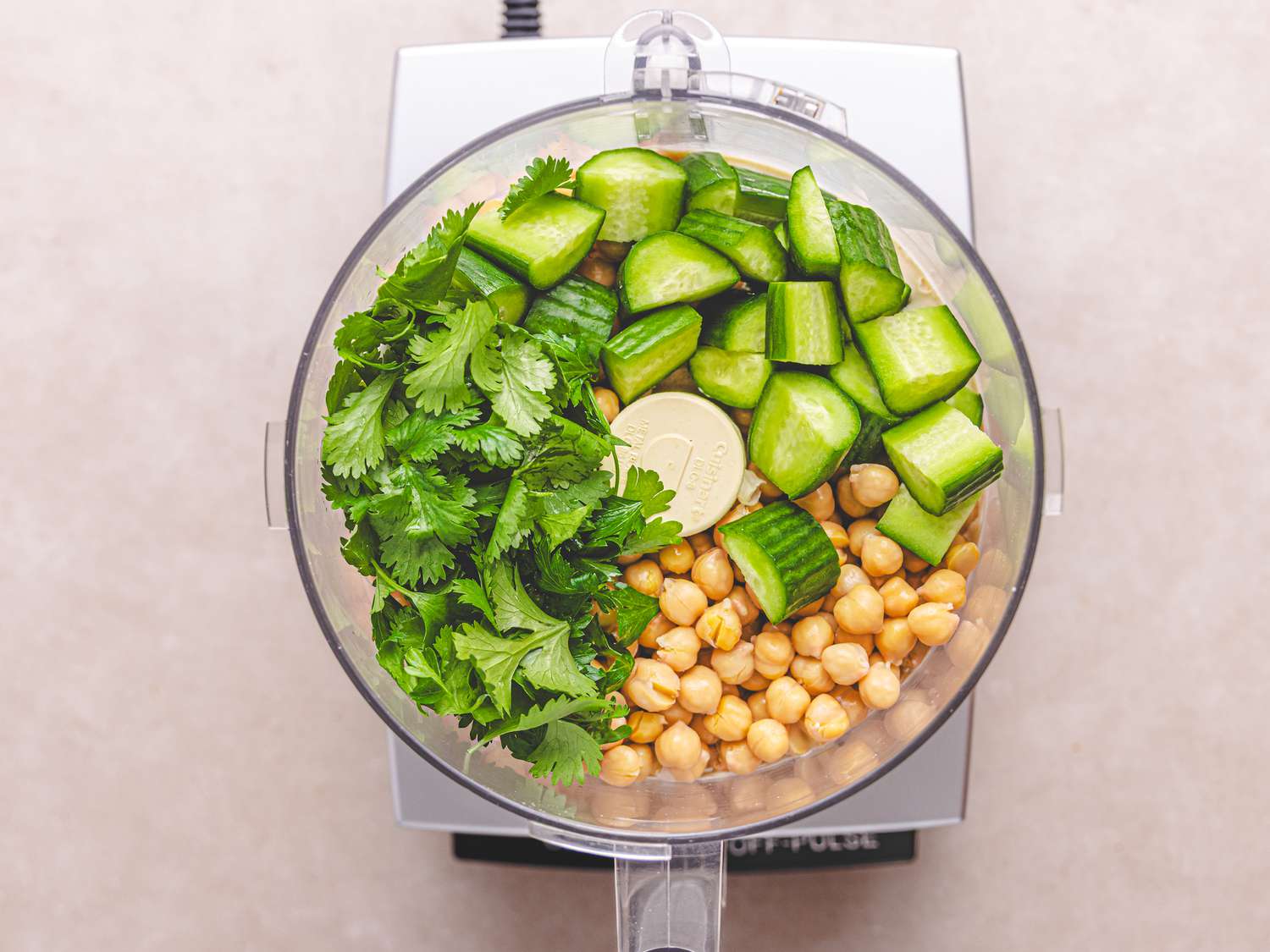 Food processor with chickpeas, chopped cucumber, cilantro, and a lime wedge inside