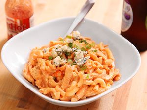 Closeup of buffalo chicken mac and cheese, served in a bowl with a sprinkle of crumbled blue cheese. A bottle of Frank's Red Hot sauce lurks in the background.