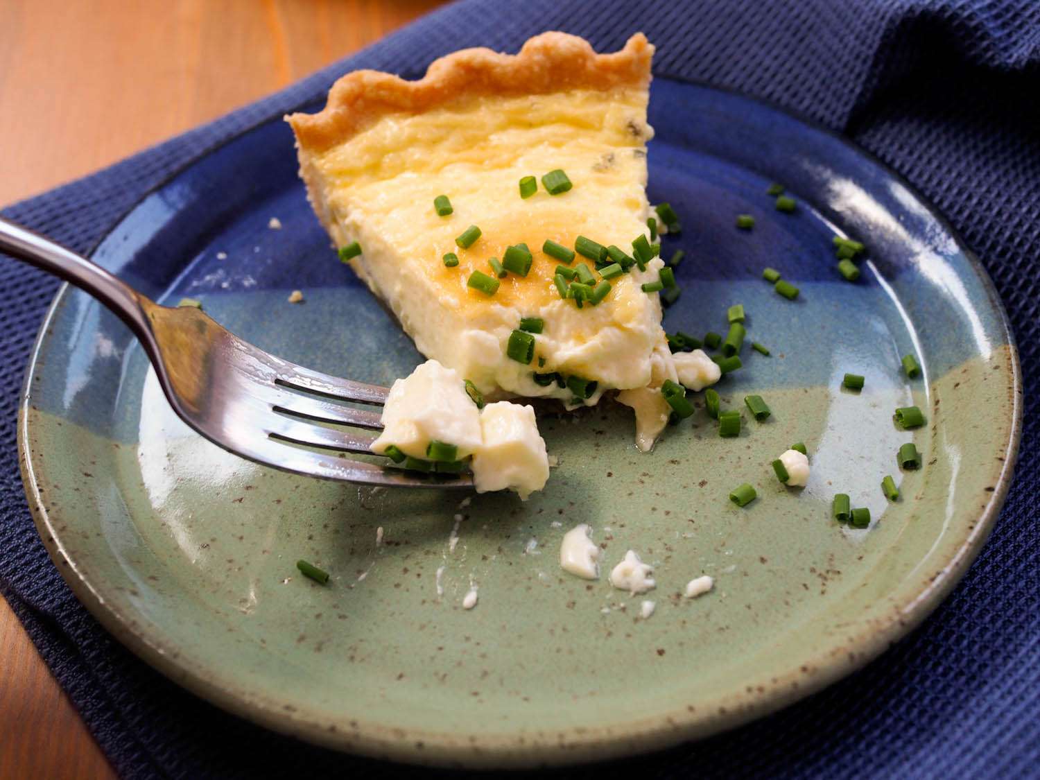 A slice of cheese quiche on a plate, sprinkled with snipped chives and partially eaten. A fork cradles a bite containing two mysterious chunks of cheese.