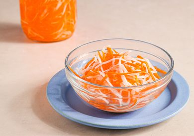 A small glass bowl on a blue plate holding Vietnamese pickled carrots and daikon. There is a jar of pickles and bring in the top lefthand corner of the image.