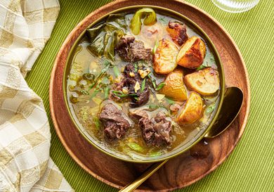 Bowl of Nilagang Beef on a wooden plate, on top of green tablecloth. 2 glasses of water, bowl of soup and napkin on the side. 