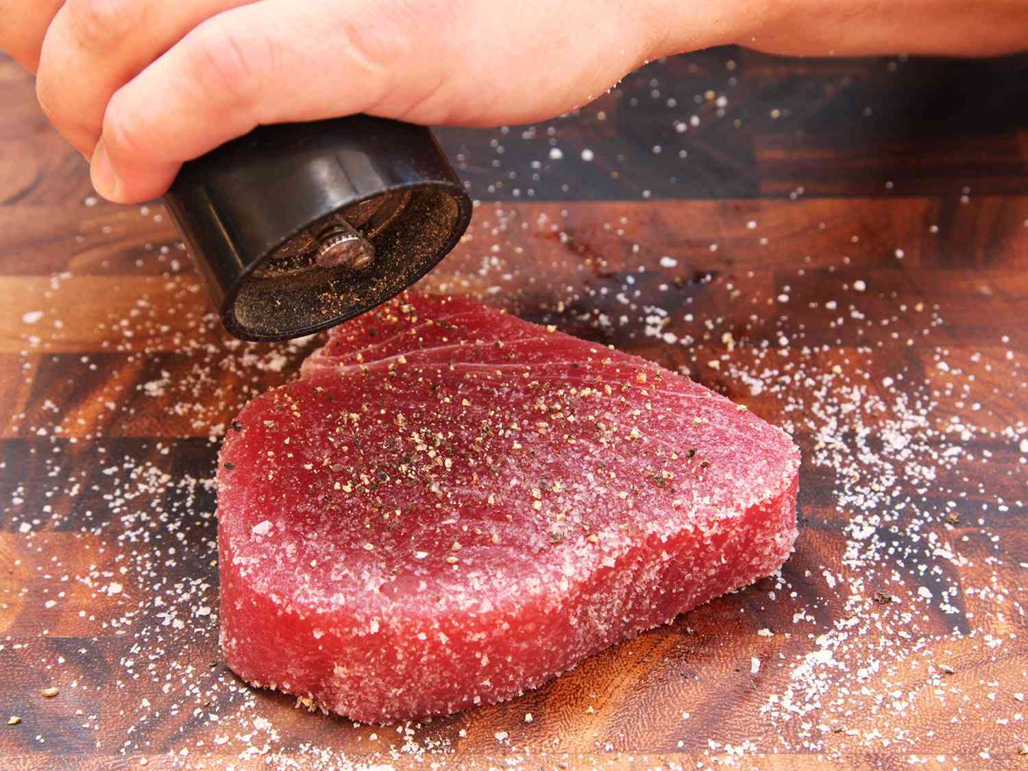 Raw tuna steak on a cutting board getting seasoned with salt and ground black pepper.