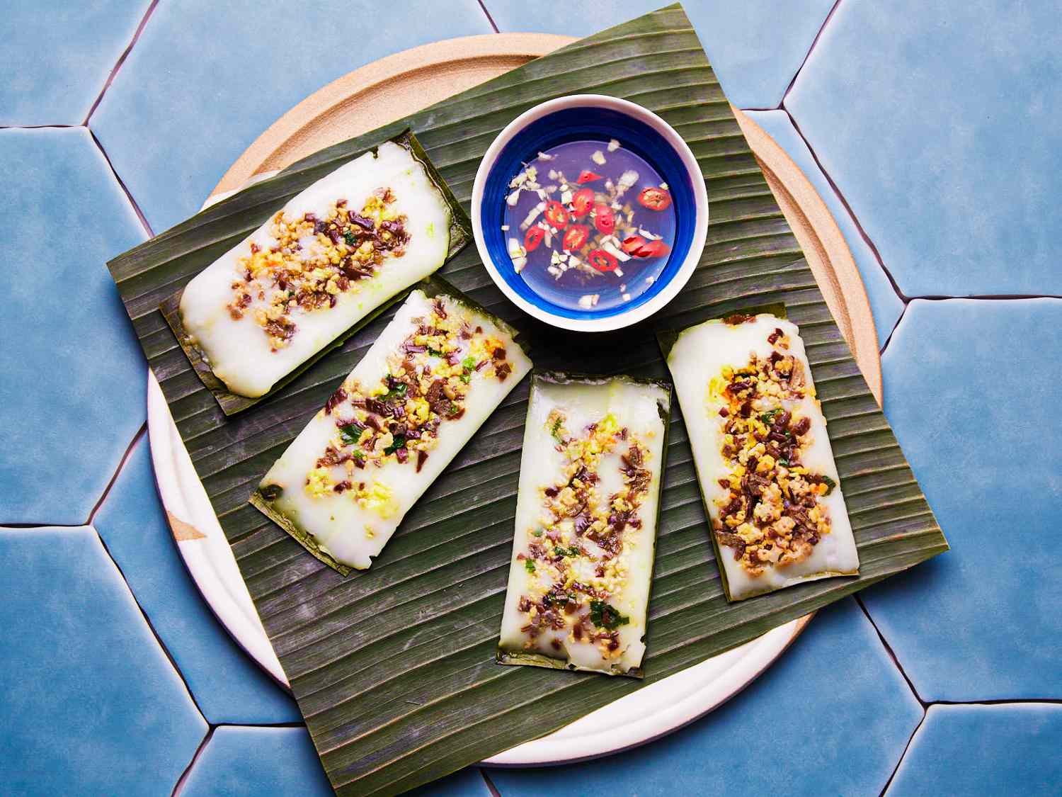 Overhead view of banh nam on a banana leaf with dipping sauce on a blue tiled background