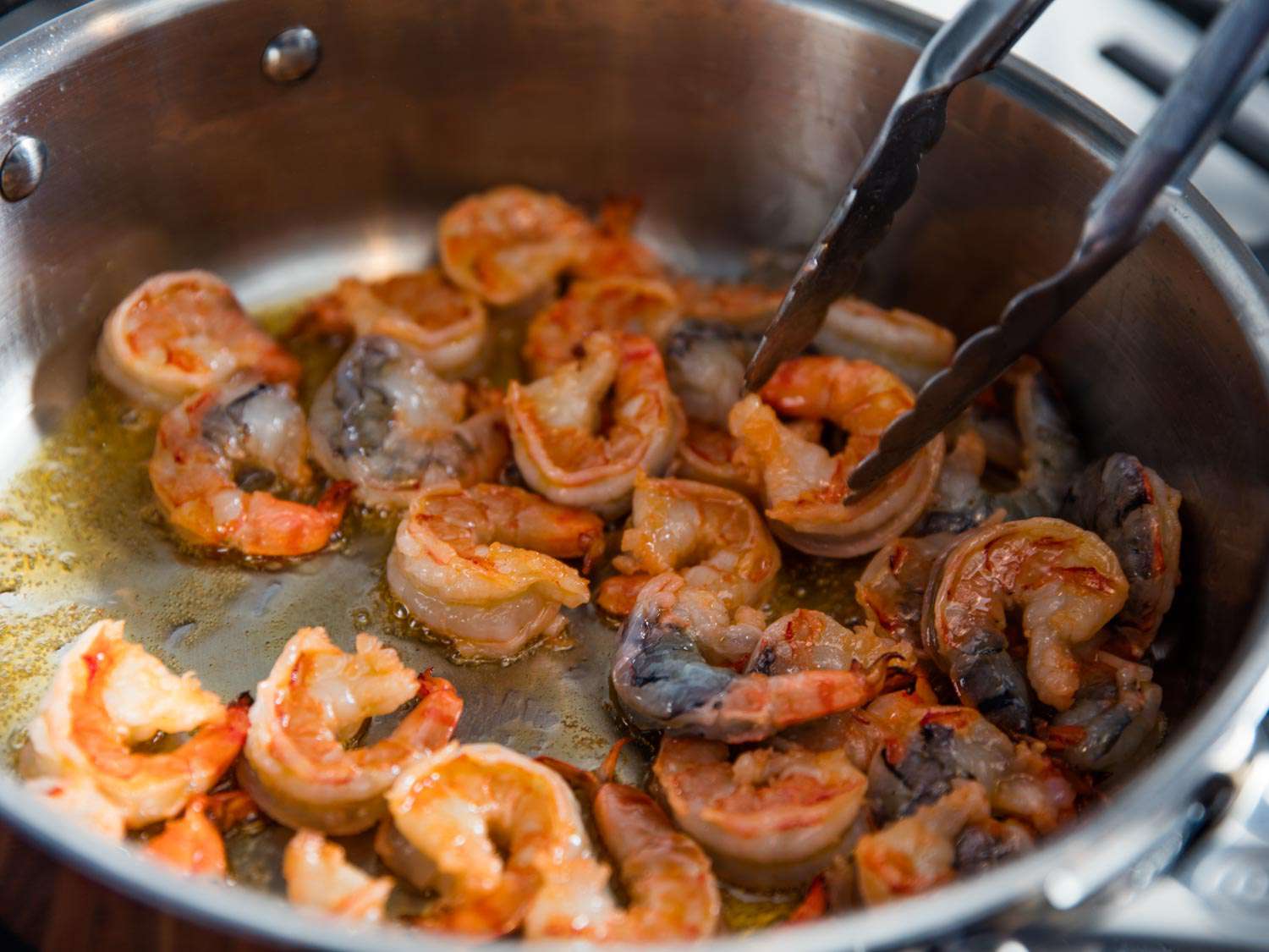 Using tongs to sauté shrimp in olive oil in a silver skillet.
