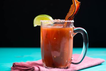 A bloody Mary in a glass mug, garnished with a strip of bacon and a lime wedge.