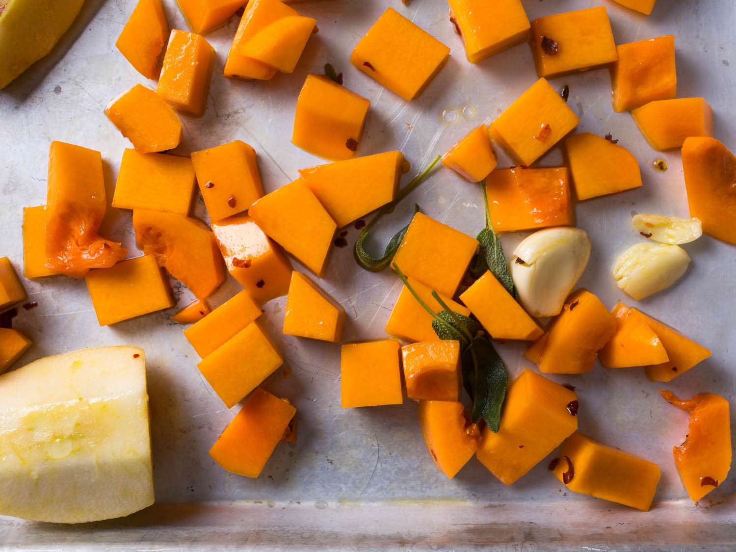 Butternut squash cubes, sage leaves, apple, and garlic tossed in olive oil on a baking sheet, preparing to be roasted. 
