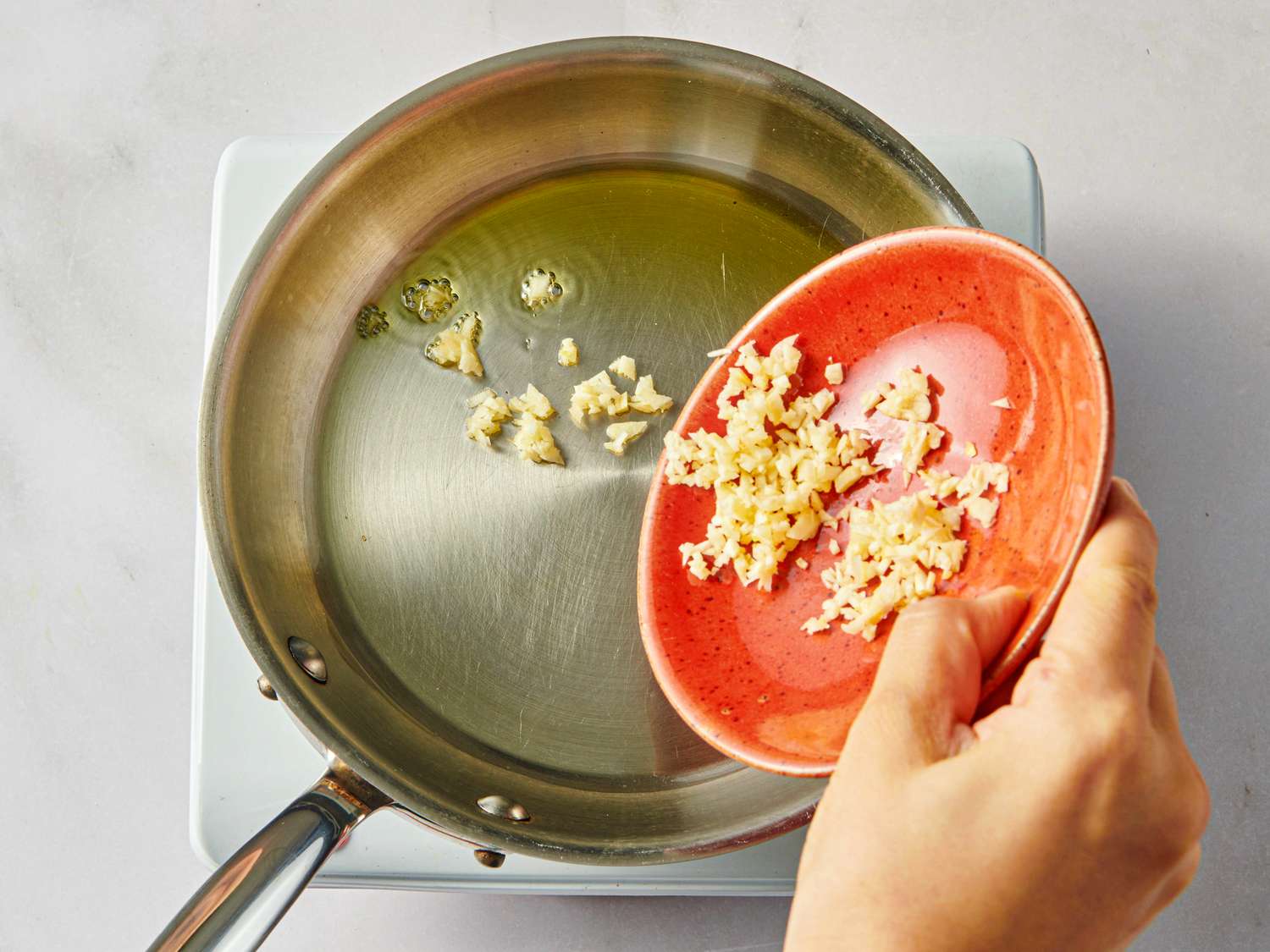 Hand adding minced garlic from an orange dish into a skillet with oil on a stovetop
