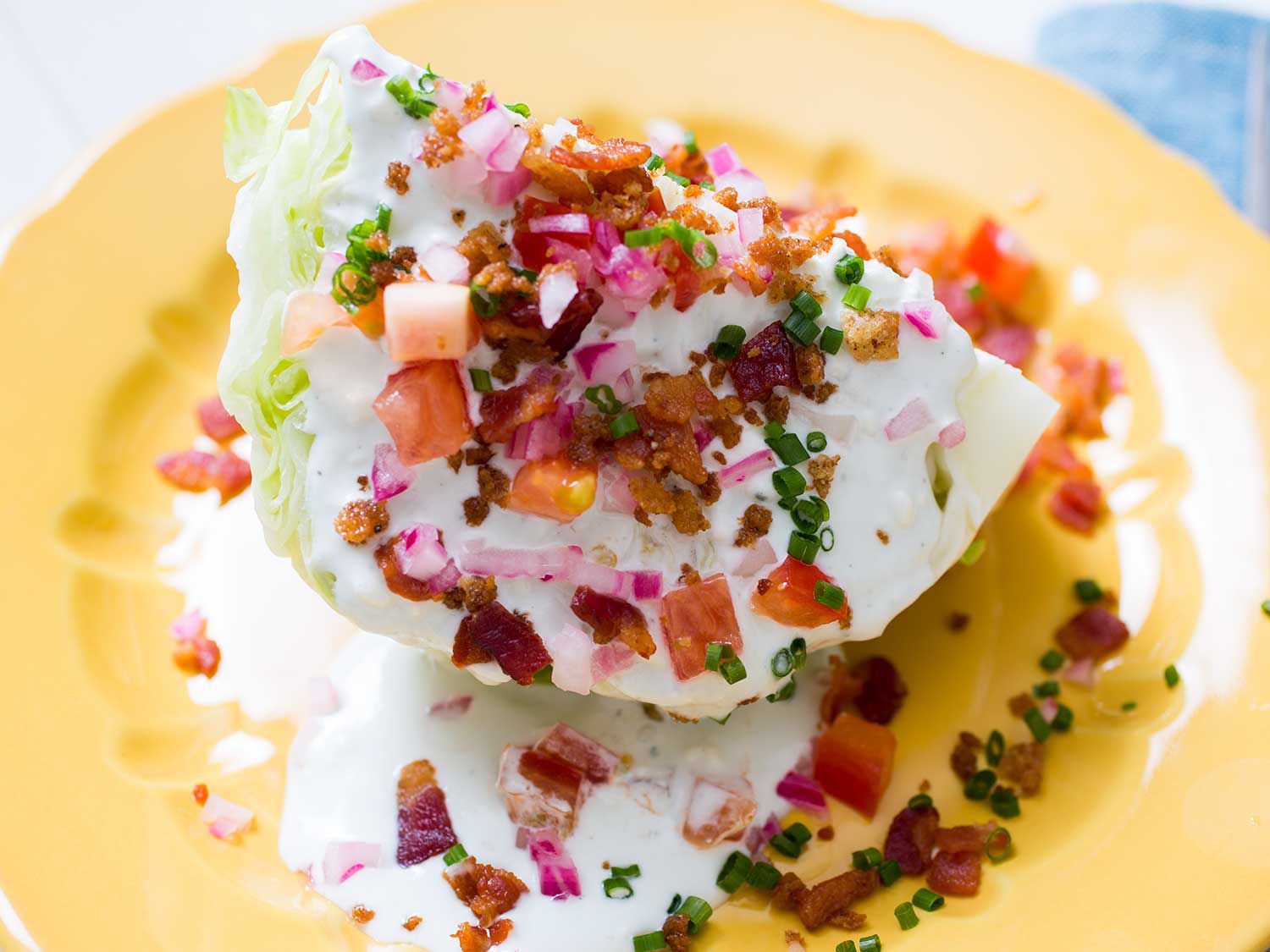 Close-up of a iceberg wedge salad drizzled with buttermilk blue cheese drssing and garnished with bacon, chives, diced tomato, and pickled red onion.