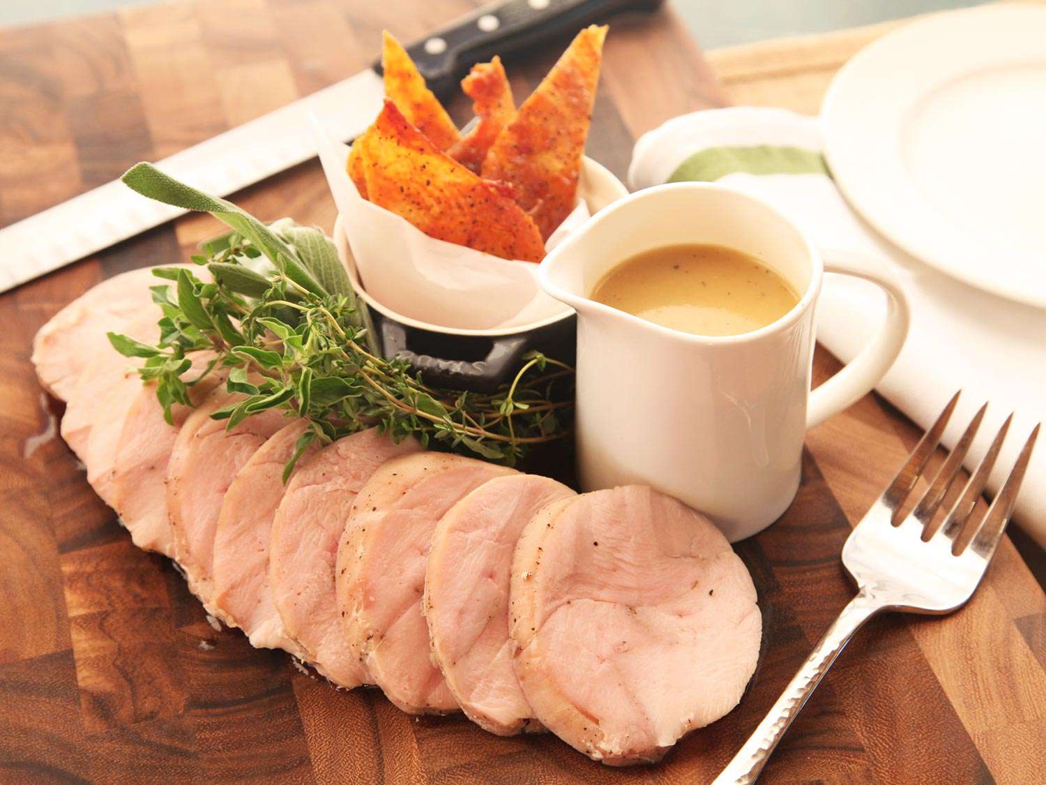 Sliced sous vide turkey breast on a wooden cutting board with herb garnish, gravy boat, and skin cracklings on the side.