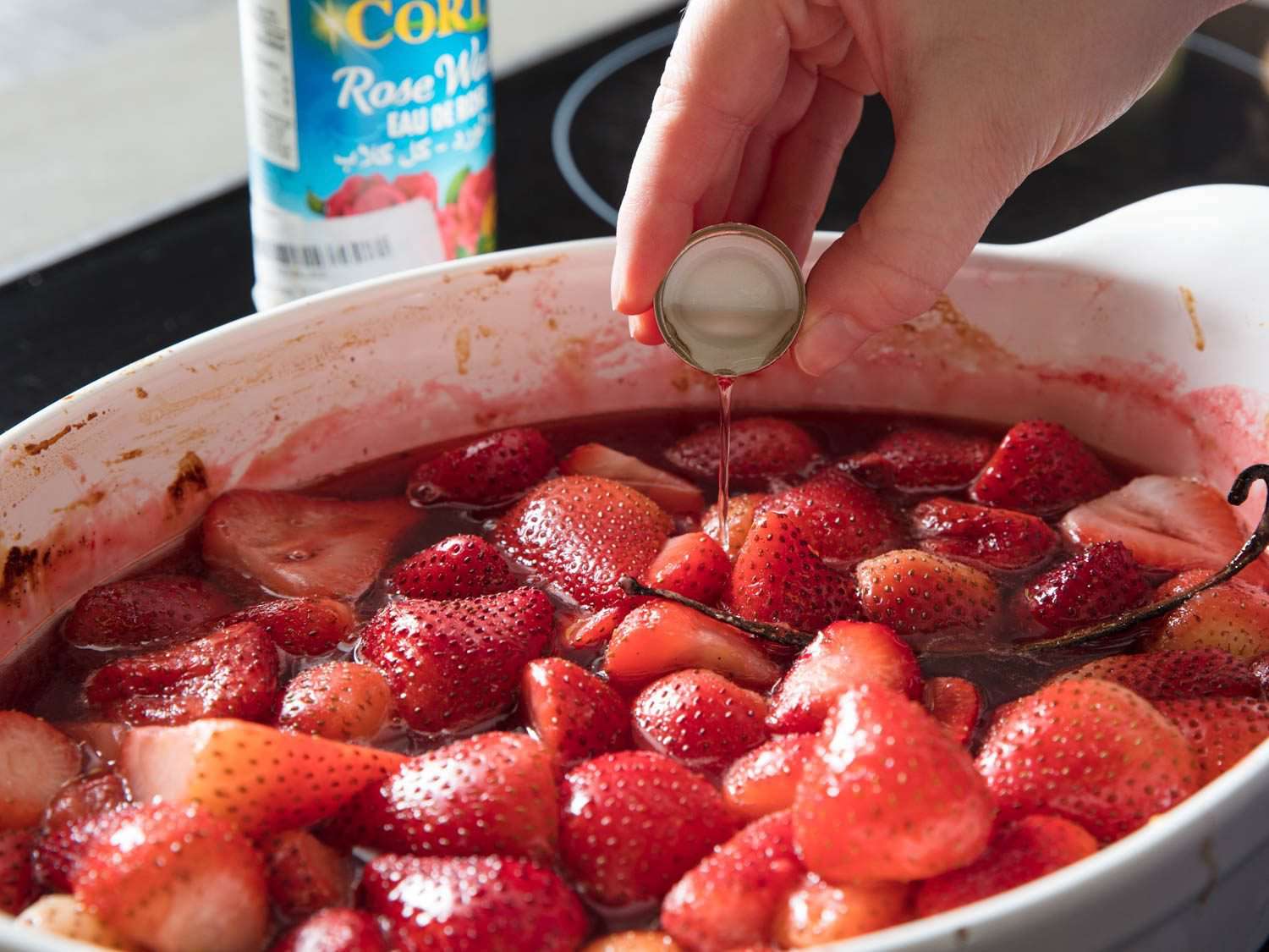 a cap-ful of rose water being tricked into a roasting dish filled with juicy sliced strawberries
