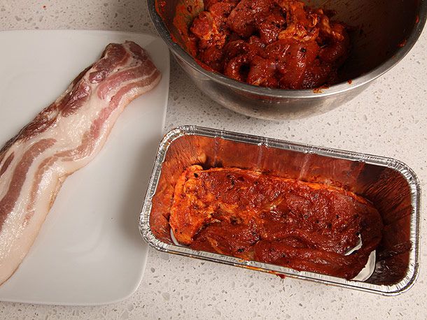 Uncooked marinated pork sirloin in aluminum loaf pan next to plate with raw bacon
