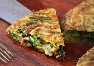 Closeup of a slice of flipped frittata with asparagus, spinach, ham, and cheese bering lifted from a cutting board with a chef's knife.