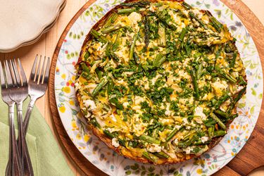 Smashed potato tart with asparagus and herbs on a floral plate, placed on a wooden table with forks and a green napkin nearby.