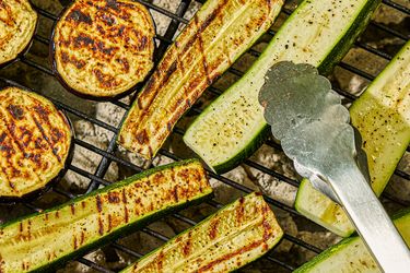 Slices of zucchini and eggplant grilling on a barbecue with metal tongs