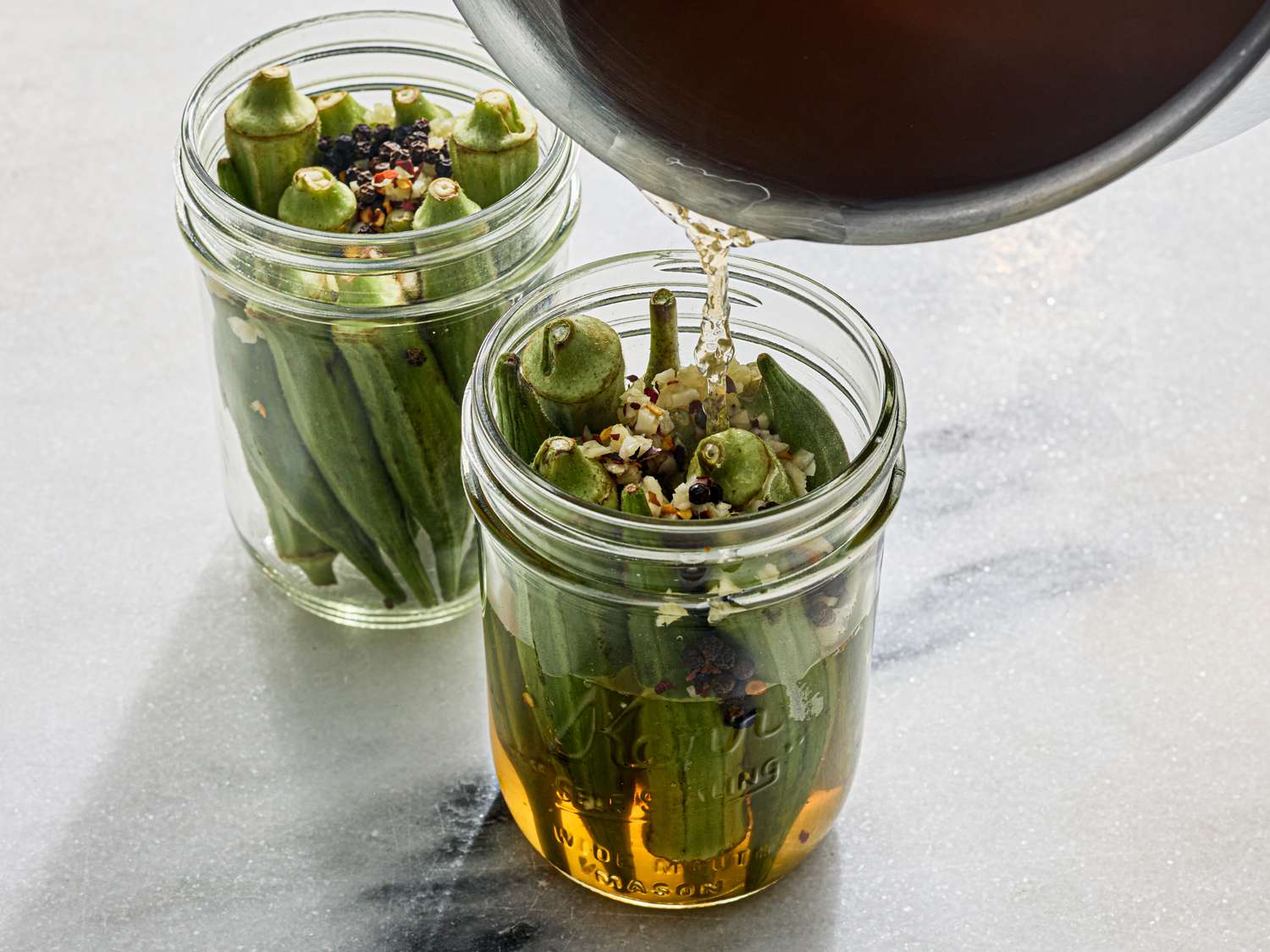 Pickling okra in glass jars with liquid being poured in