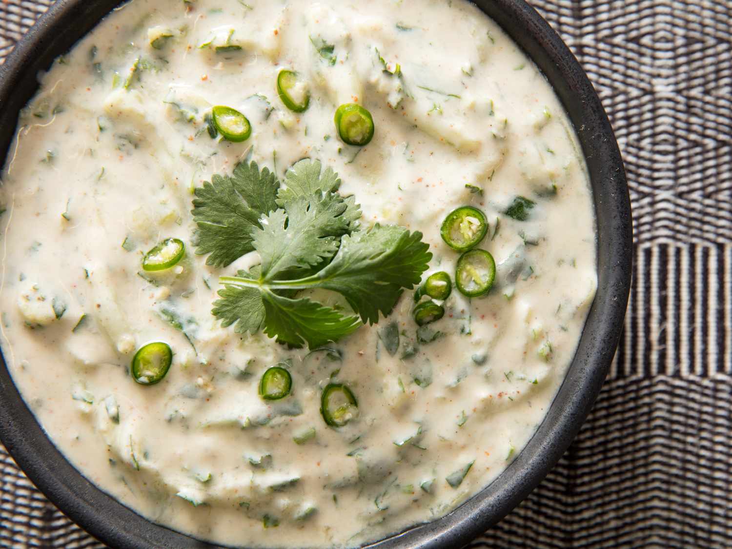 Overhead closeup of cucumber raita, served in a black bowl and garnished with sliced serrano chile and a sprig of cilantro.