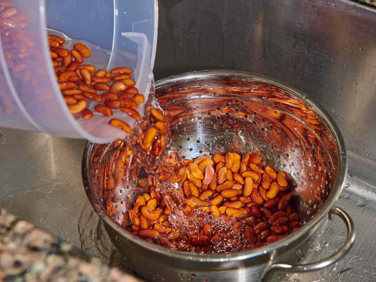 Soaked kidney beans being drained in a metal colander inside of a deep stainless steel-lined sink.