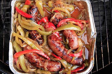 Overhead view of a finished batch of grilled Italian sausage with peppers and onions, piled in a disposable aluminum pan on a grill.