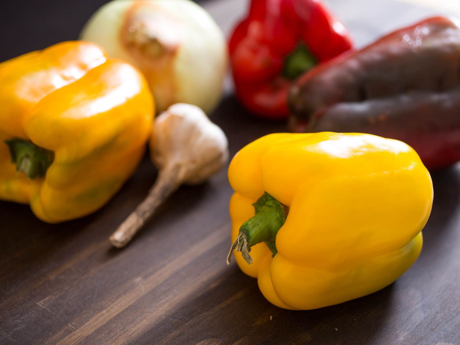 Red bell pepper, yellow bell pepper, garlic, and onion resting on a wooden surface.