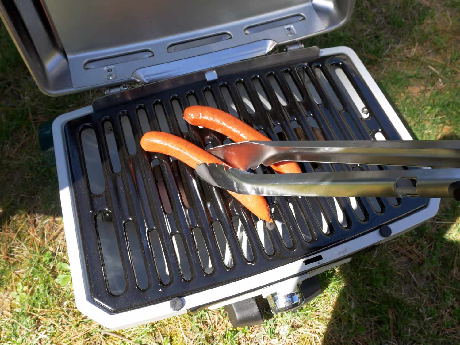 a person using a pair of grill tongs to flip a hot dog on a portable gas grill