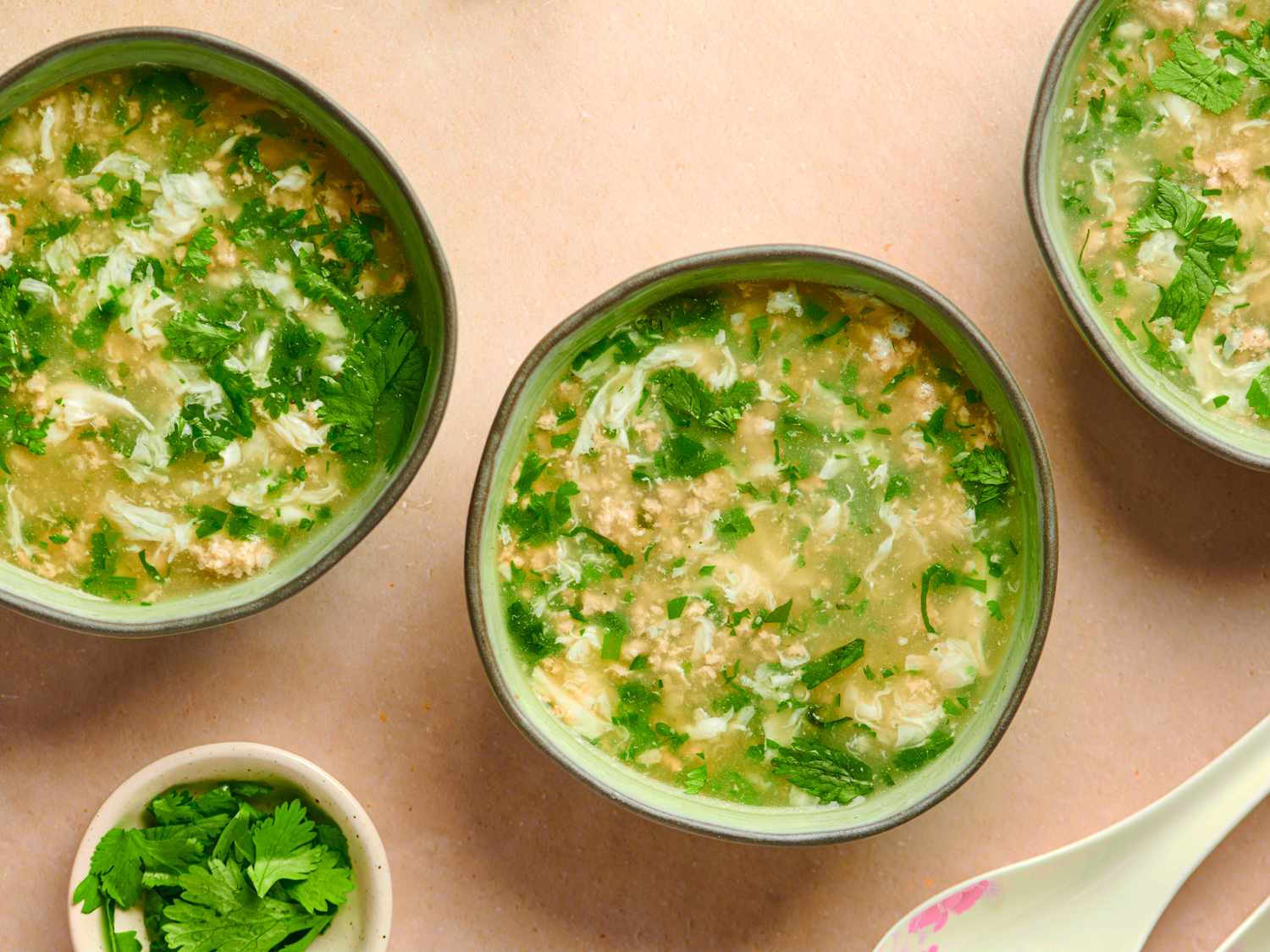 Three bowls of West Lake Soup on a pink hued counter. There is a soup spoon in the bottom right corner of the image, and a small bowl of cilantro leaves in the bottom left corner.