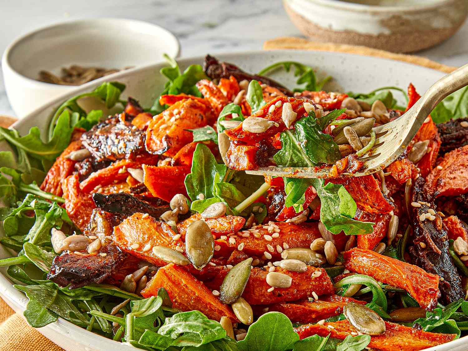 A roasted carrot and date salad with seeds on a bed of greens served in a wide bowl with a fork