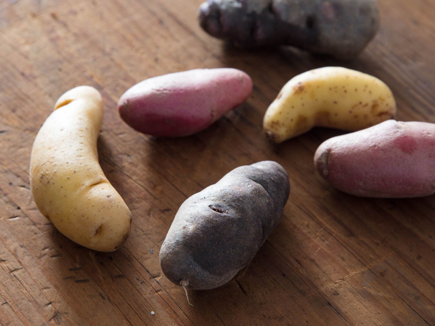 Six fingerling potatoes in various colors on a wooden cutting board.