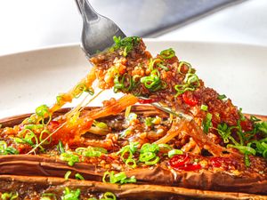 A fork lifting a piece of Chinesestyle BBQ eggplant garnished with chopped green onions on a plate