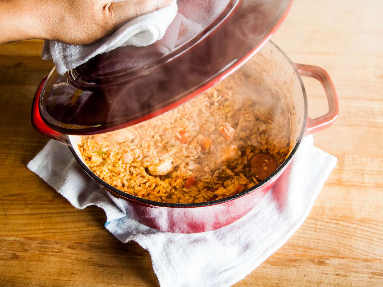 Hand lifting the lid of a Dutch oven containing steaming jambalaya