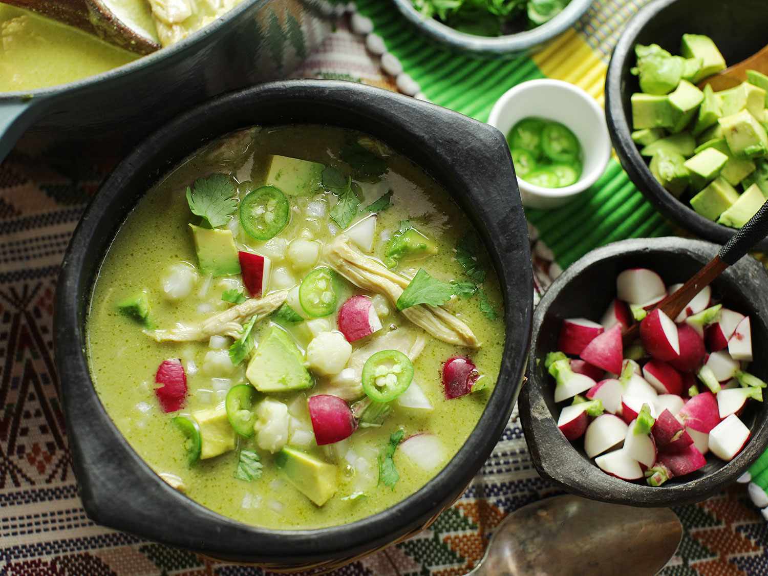 Overhead view of green posole, served in an earthenware bowl with sides of diced radish and avocado for garnishing.