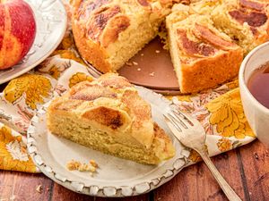 A slice of apple cinnamon cake on a plate part of the cake and an apple shown on a decorative table setting