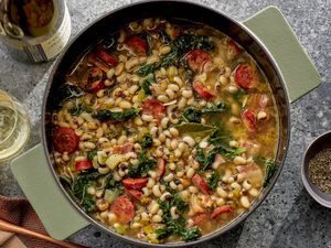 Pot of black eyed peas, kale and sausage stew, surrounded by a printed napkin, bottle and glass of wine on a stone surface 