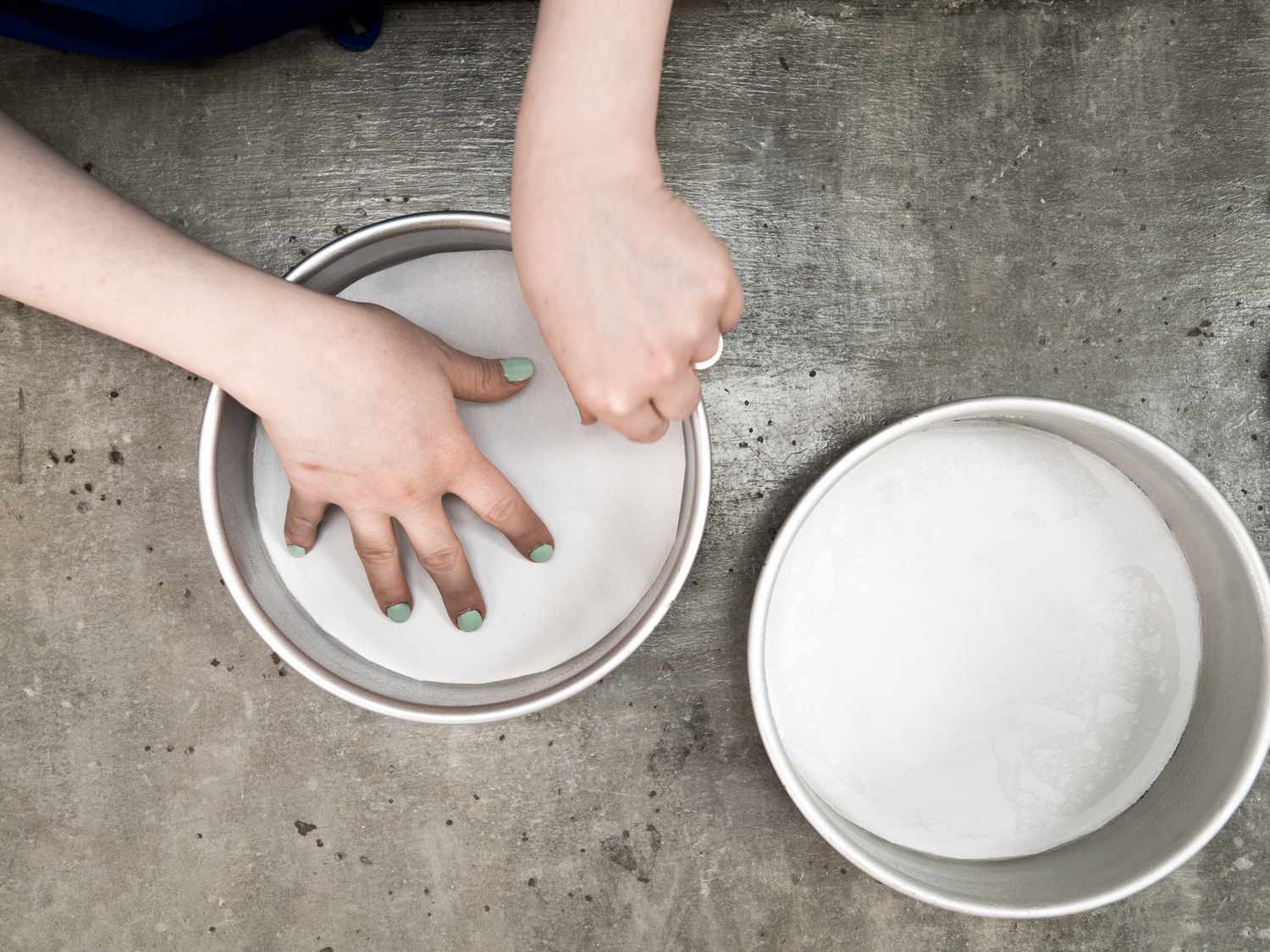 One round cake pan lined with a circle of parchment, hands pressing a circle of parchment into a second cake pan.