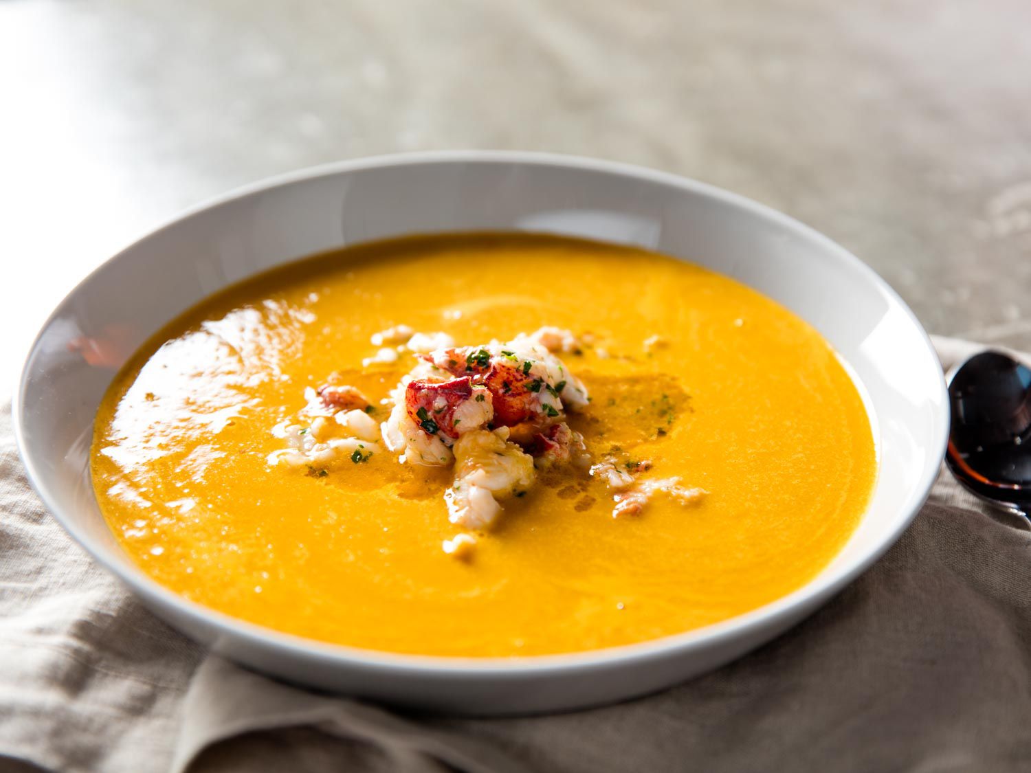 A serving of the finished bisque in a shallow white bowl, a large spoonful of lobster meat is mounded in the center, drizzled with the herb-butter mixture it was sautéed in.