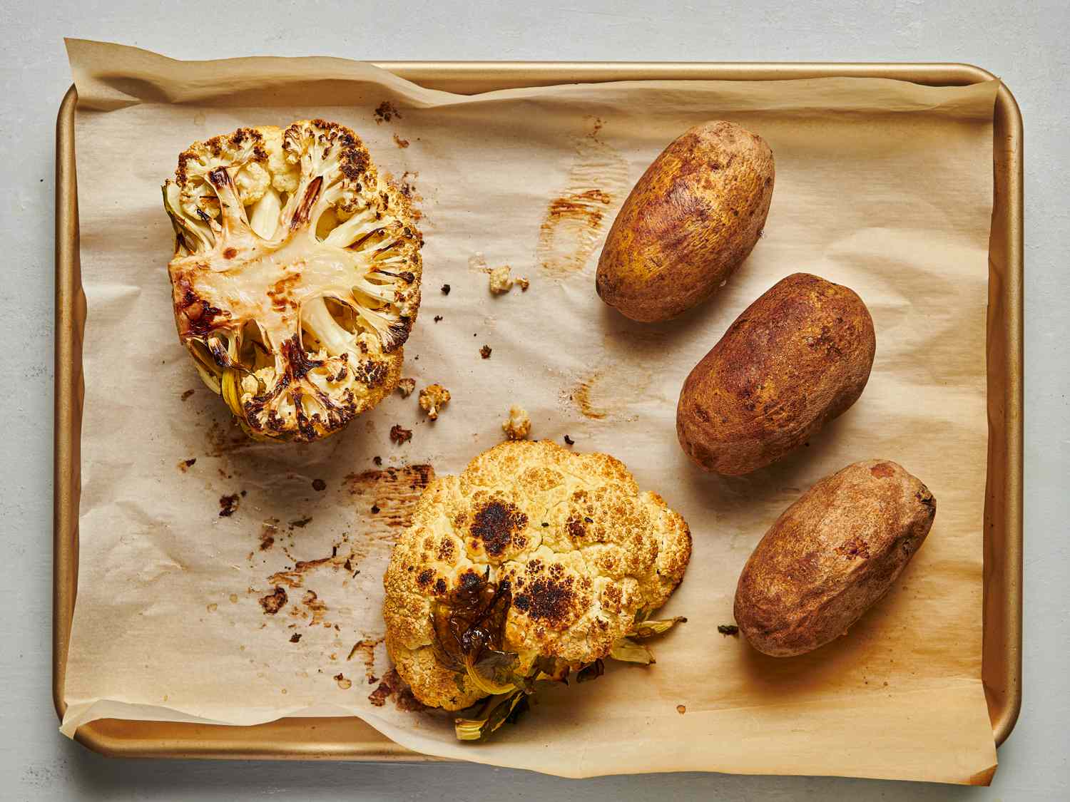 A baking sheet lined with parchment paper, holding roasted vegetables. There is an entire cauliflower, cut in half, and three whole russet potatoes.