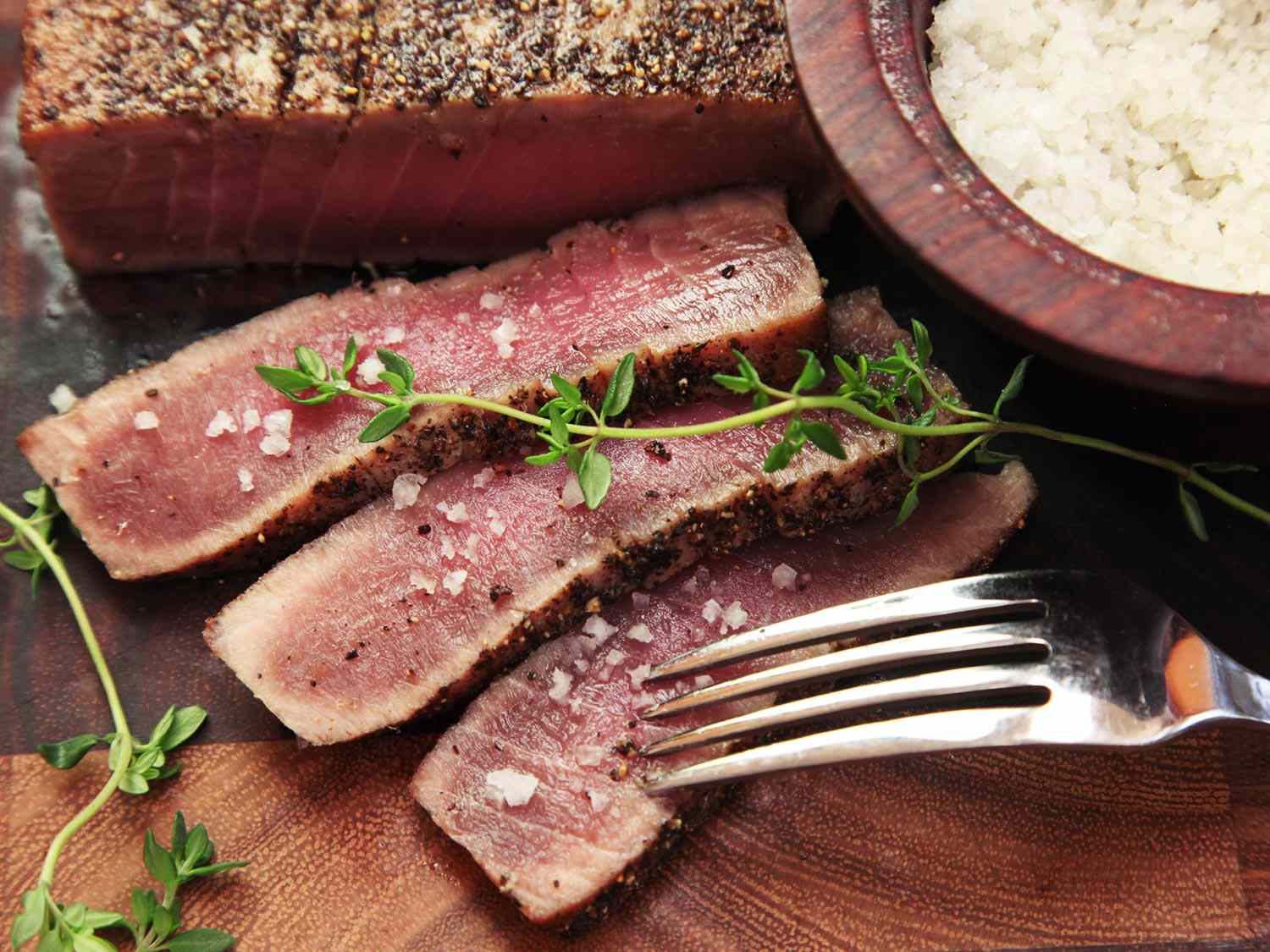 Close-up of the seared tuna steak on a cutting board. Several slices of the tuna have been shingled beside it, revealing a moist, translucent center. Coarse salt has been lightly sprinkled over the slices (a bowl of salt sits to one side of the tuna). Two sprigs of thyme are strewn over the slices and a fork is about to skewer the bottom slice and abscond with it.