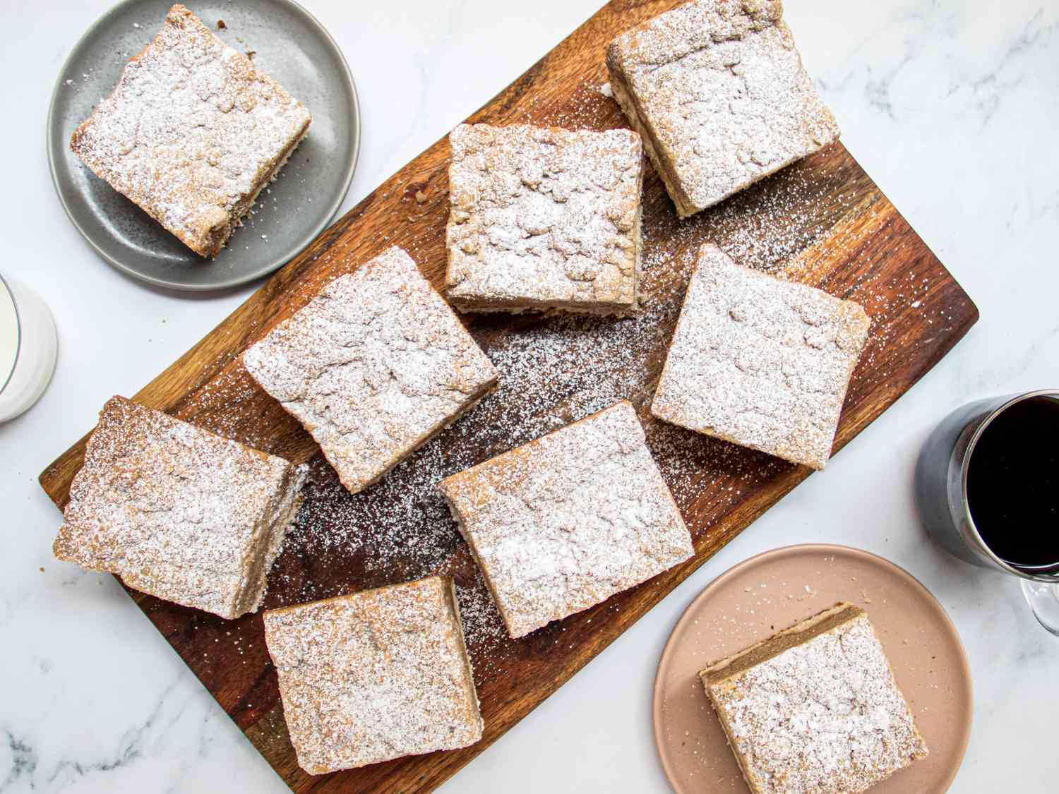 NJ Crumb Buns dusted with sugar on a wooden board. 2 small dishes have crumb bun squares on them, with cups of milk and coffee