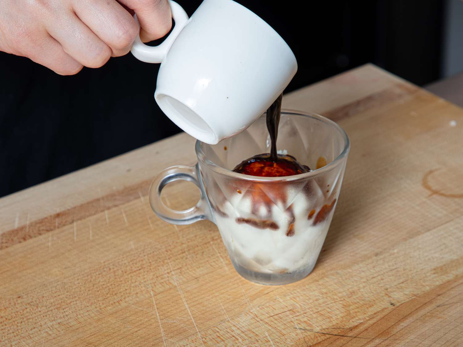 Hand pouring espresso over ice cream in a glass on a wooden surface