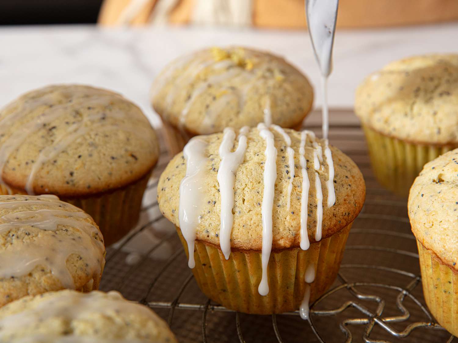 Lemon poppy seed muffins on a cooling rack, with glaze being drizzled on top of one muffin
