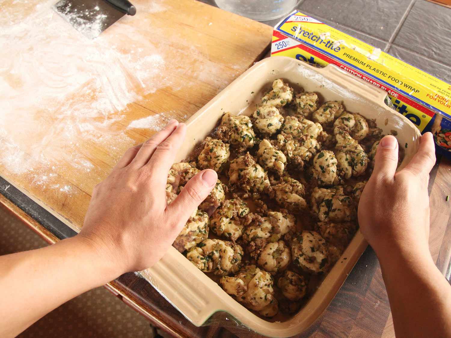 Hands framing a baking dish full of unbaked pull-apart stuffing rolls, next to a box of plastic wrap