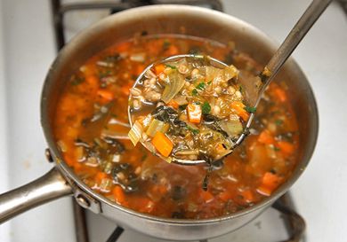 A ladle full of vegetarian escarole and parmesan soup is held up for the camera. A saucepan of the soup is visible in the background.