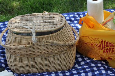 A wicker picnic basket on a blue checkered tablecloth spread on the grass outdoors. There is a Serious Eats bag with paper towels and other picnic supplies next to the basket. 