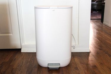 A white trash bin in a room with wooden flooring and a partial view of a door and furniture in the background