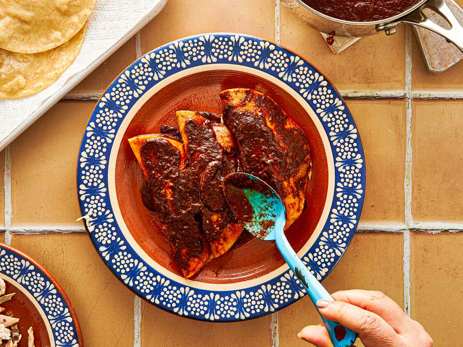 A person spooning sauce over enchiladas in a decorative ceramic dish on a tiled surface with tortillas and a pot nearby