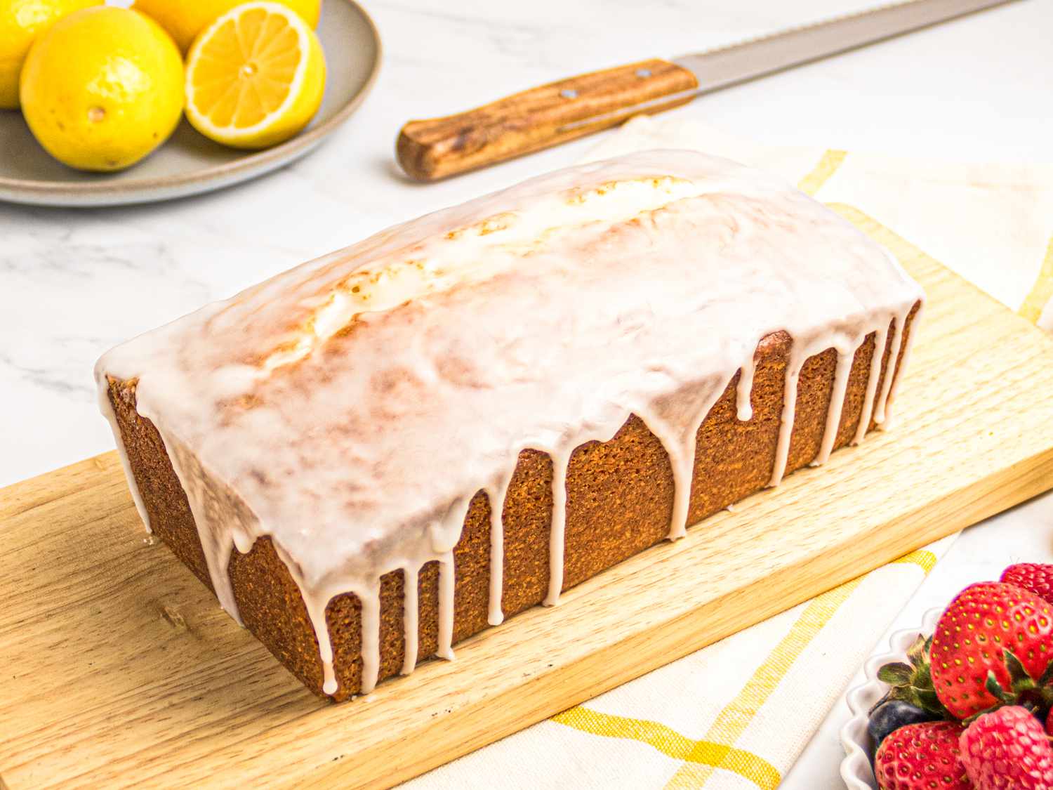 Lemon pound cake with glaze on a wooden board, surrounded by lemons, a knife, and a bowl of fresh berries.