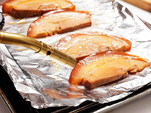 A blow torch is crisping up chashu pork slices on a foil lined baking sheet. 