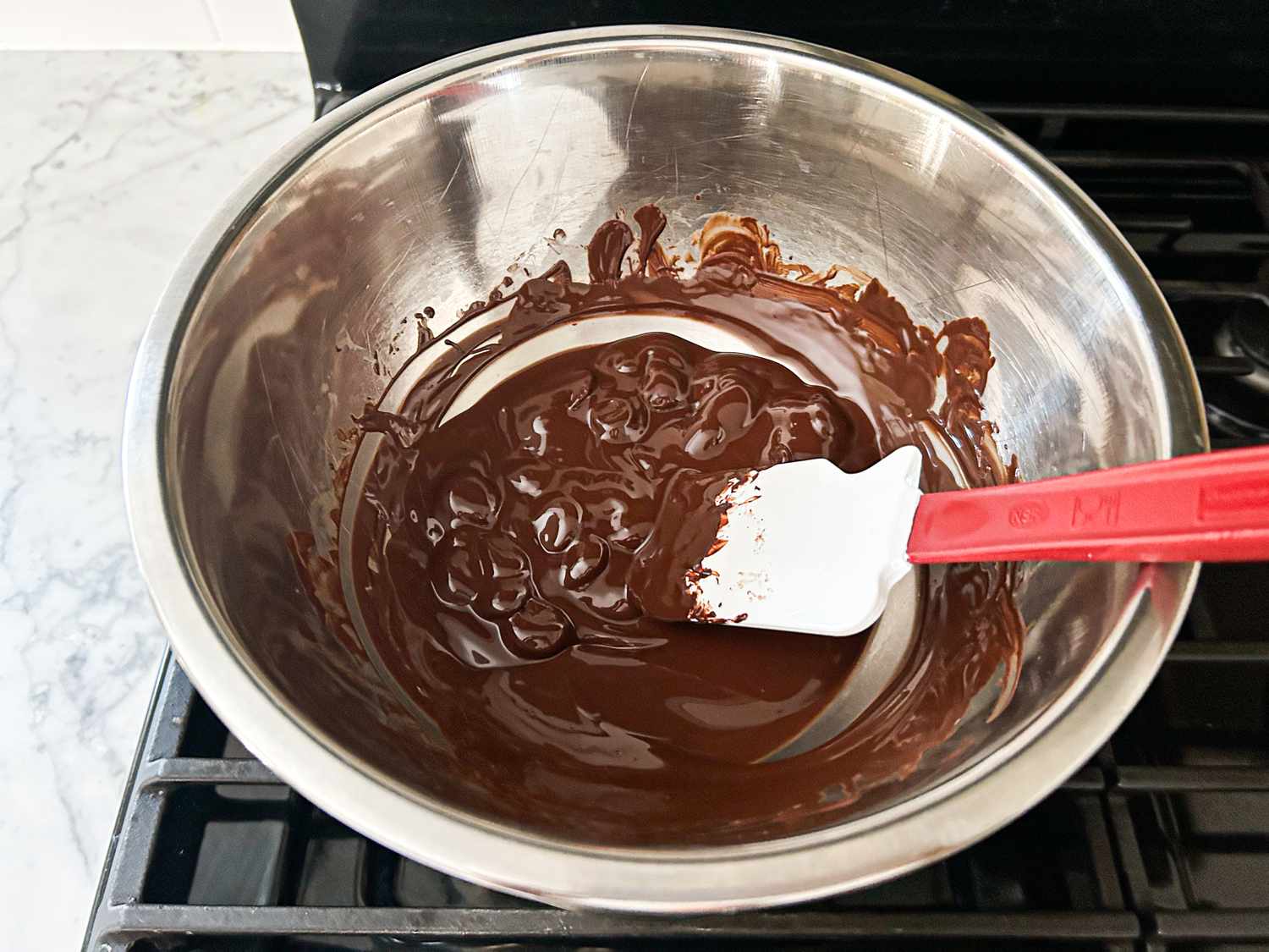 A bowl of melted chocolate being stirred with a red spatula on a stovetop