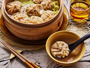 Angled view of a tablescape with a bamboo steamer full of dumplings behind a biten into soup dumpling on a soup spoon in an individual bowl on a black and white marbled surface. Overall tone and lighting is warm
